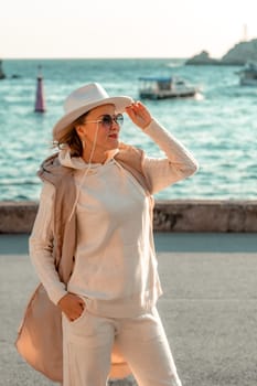 Happy blonde woman in a white suit and hat posing at the camera against the backdrop of the sea.