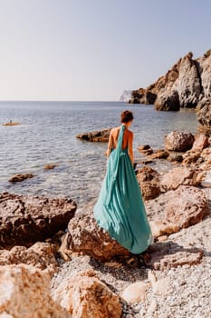 Woman green dress sea. Woman in a long mint dress posing on a beach with rocks on sunny day. Girl on the nature on blue sky background