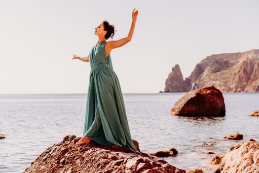 Woman green dress sea. Woman in a long mint dress posing on a beach with rocks on sunny day. Girl on the nature on blue sky background