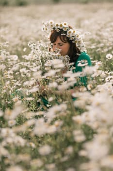 Happy woman in a field of daisies with a wreath of wildflowers on her head. woman in a green dress in a field of white flowers. Charming woman with a bouquet of daisies, tender summer photo.