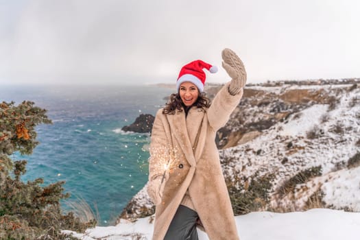 Outdoor winter portrait of happy smiling woman, light faux fur coat holding heart sparkler, posing against sea and snow background.