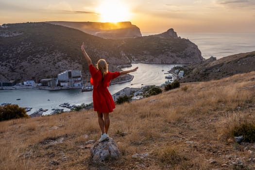 Happy woman standing with her back on the sunset in nature in summer with open hands posing with mountains on sunset, silhouette. Woman in the mountains red dress, eco friendly, summer landscape active rest.