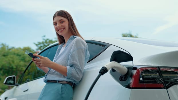 Young woman recharging battery for electric car during road trip travel EV car in natural forest or national park. Eco friendly travel during vacation and holiday. Exalt