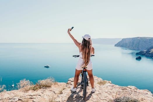 A woman cyclist on a mountain bike looking at the landscape of mountains and sea. Rear view of cyclist woman standing in front to the sea with outstretched arms. Freedom and healthy lifestyle concept