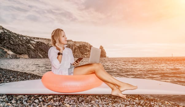 Successful business woman in yellow hat working on laptop by the sea. Pretty lady typing on computer at summer day outdoors. Freelance, travel and holidays concept.