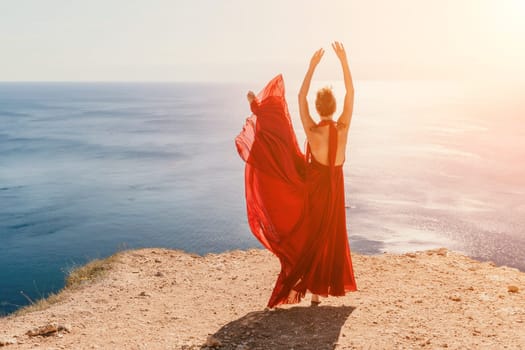 Side view a Young beautiful sensual woman in a red long dress posing on a rock high above the sea during sunrise. Girl on the nature on blue sky background. Fashion photo.