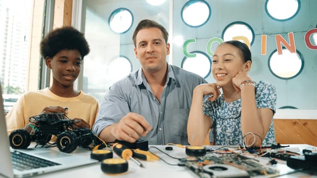 Teacher explain about electronic equipment while student with mixed races fixing car model at table with laptop and electric tool. Diverse woman learning about robotic machine structure. Edification.