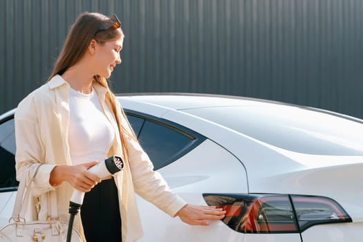 Young woman holding shopping bag recharge EV car battery from charging station at parking lot. Modern woman go shopping by environmental friendly electric vehicle in urban travel lifestyle. Expedient