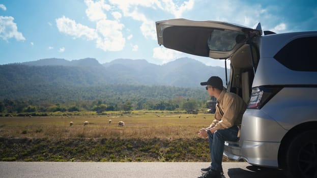 Man sitting in open trunk on a country road and enjoying a beautiful mountain view.