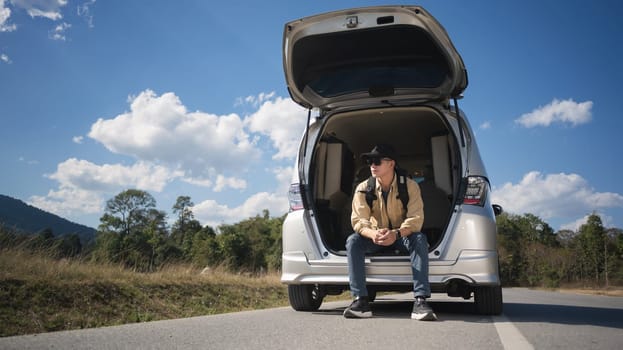 Male traveler on a road trip, sitting on the open trunk of his car against blue sky and white cloud.