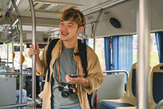 Smiling Asian man with backpack standing in bus and traveling with public transport.