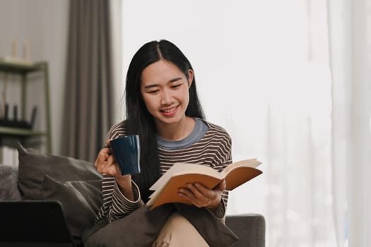 Pleased young woman in casual clothes drinking coffee and reading book on couch.