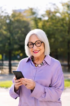 senior woman looking at camera smiling happy using mobile phone outdoors, concept of technology and elderly people leisure, copy space for text