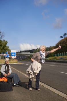 Women, road and hitch hiker for travel in retirement, lift and friends in city with bags for adventure. Senior ladies, thumbs up and happy by roadside with hand gesture and stop transport to commute.