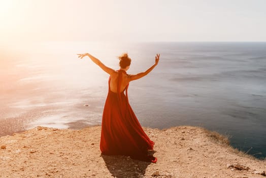 Side view a Young beautiful sensual woman in a red long dress posing on a rock high above the sea during sunrise. Girl on the nature on blue sky background. Fashion photo.