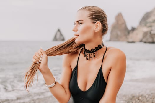 Woman travel portrait. Happy woman with long hair poses on a red volcanic rock at the beach. Close up portrait cute woman in black bikini, smiles at the camera, with the sea in the background