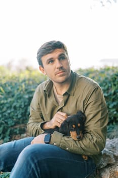 Young man sits in a park and strokes a black puppy on his knees while looking away. High quality photo