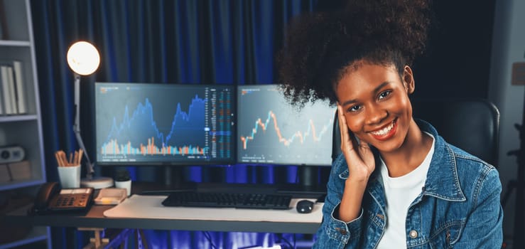 Profile of young African American businesswoman smiling on happy face wearing jeans shirt, sitting on chair against stock exchange market screen background. Concept of investment blogger. Tastemaker.