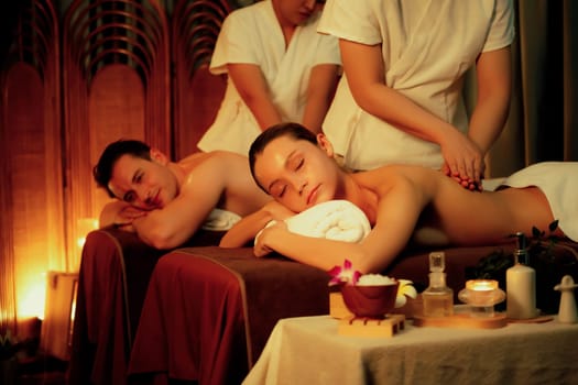 Caucasian couple customer enjoying relaxing anti-stress spa massage and pampering with beauty skin recreation leisure in warm candle lighting ambient salon spa at luxury resort or hotel. Quiescent