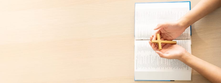 Female god believer holding wooden cross on opened holy bible book at light wooden church table. Top view. Concept of hope, religion, faith, christianity and god blessing. Warm background. Burgeoning.