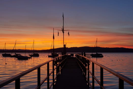 Bodensee Lake Sunrise Panorama. Morning Sunlight Over Tranquil Waters. Witness the mesmerizing dawn over Germany's Bodensee Lake, captured from a boat dock. Embrace the tranquil beauty of the early morning as the sun rises, casting a soft glow on the landscape. The peaceful scene features boats, yachts, and a charming water shack set against a backdrop of a captivating sky. Clouds delicately reflect on the calm water, creating a serene atmosphere. Immerse yourself in the serene beauty of a lakeside sunrise. Explore the harmony of nature, technology, and production as the day unfolds by the lake.