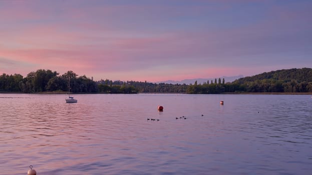 Bodensee Lake Sunrise Panorama. Morning Sunlight Over Tranquil Waters. Witness the mesmerizing dawn over Germany's Bodensee Lake, captured from a boat dock. Embrace the tranquil beauty of the early morning as the sun rises, casting a soft glow on the landscape. The peaceful scene features boats, yachts, and a charming water shack set against a backdrop of a captivating sky. Clouds delicately reflect on the calm water, creating a serene atmosphere. Immerse yourself in the serene beauty of a lakeside sunrise. Explore the harmony of nature, technology, and production as the day unfolds by the lake.