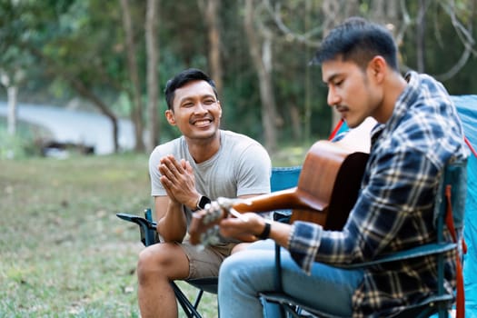 LGBTQIA Gay couple camping together in woods for holidays and relax on guitar together.