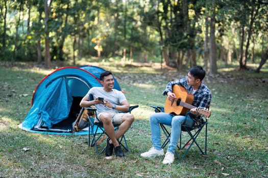 LGBTQIA Gay couple camping together in woods for holidays and relax on guitar together.