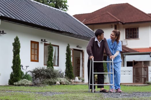 Nurse or caregiver helps elderly walk by using walker in garden.