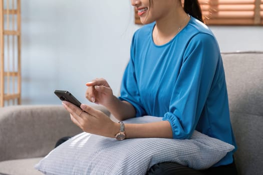 Happy young Asian woman using mobile phone at home, messaging or browsing social networks while relaxing on sofa.