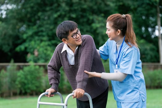 Nurse or caregiver helps elderly walk by using walker in garden.