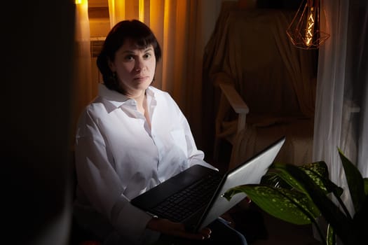 A girl in white shirt with a laptop in a dark room. A middle-aged woman works online remotely in the evening. The student is preparing for the exam at night. Blogger or freelancer in the chat