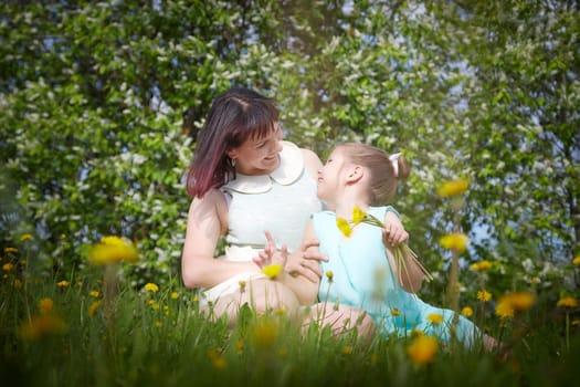 Happy mother and daughter enjoying rest, playing and fun on nature on a green lawn with dandelions and blooming apple tree on background. Woman and girl resting outdoors in summer and spring day