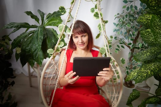 A girl with a laptop and notebook is working on a swing chair in the winter garden. A middle-aged business woman. Psychologist in the office with flowers. Online communication and consultation