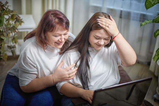 Fat funny funny adult mother and daughter with laptop indoors. A teenage girl teaches a middle-aged woman modern technology. Internet, chatting