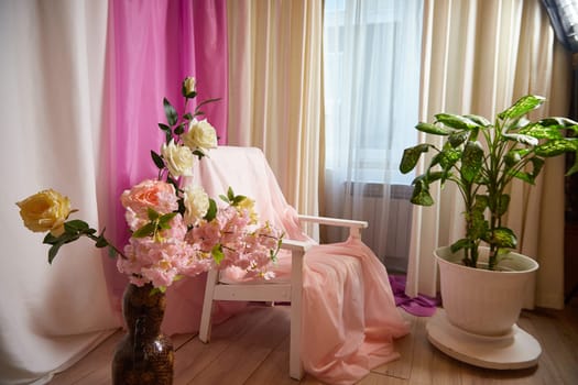 Delicate pink bright interior of the room with an armchair, vase with roses, draped curtains and a window. Location and background in the photo studio