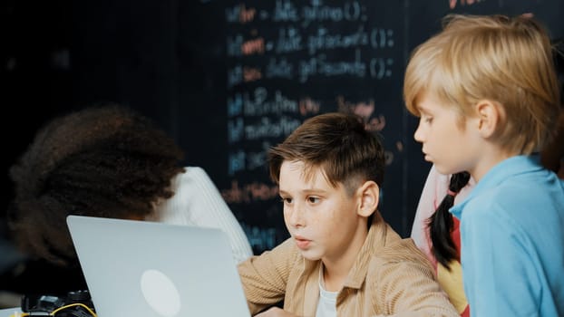 Closeup of boy using laptop programing engineering code and writing program while group of smart diverse student standing surrounded by friend in STEM technology classroom at blackboard. Erudition.