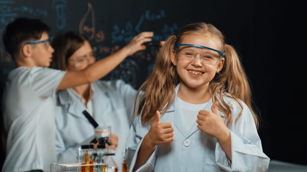 Smart caucasian girl showing thumb while teacher and student doing experiment behind. Child looking at camera while boy looking under microscope at black board written chemical theory. Erudition.