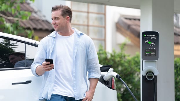 Modern eco-friendly man recharging electric vehicle from home EV charging station. EV technology utilization for tracking energy usage to optimize battery charging at home. Panorama Synchronos