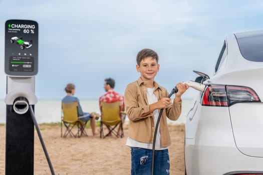 Family vacation trip traveling by the beach with electric car, little boy recharge EV car while his family enjoy seascape beach. Family trip with alternative energy and eco-friendly car. Perpetual