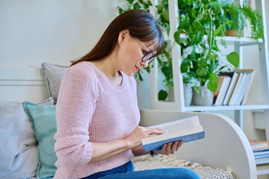 Mature woman reading paper book sitting on couch at home. Relaxation, literature hobby, fiction, lifestyle, people concept