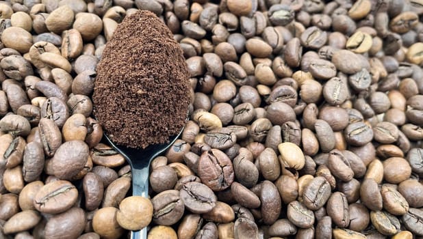 Against the background of roasted aromatic coffee beans lies a metal spoon filled with ground coffee. A drink made from roasted and ground beans from the coffee tree or coffee bush