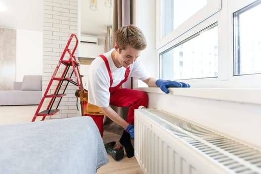 Young handsome professional plumber worker installing heating radiator in an empty room of a newly built apartment or house. Construction, maintenance and repair concept.