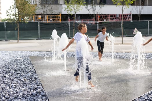 Cheerful young teen girl in city fountain, girl in wet clothes is having fun and enjoying the cool summer water, background city architecture. High quality photo