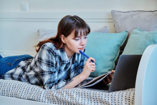 College student girl studying at home, lying on the couch with laptop, making notes in study book. Education, training, technology, youth concept