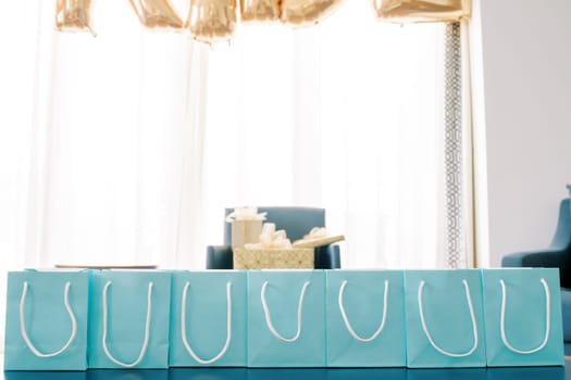 Row of blue gift bags with white handles stands on a table in the room. High quality photo