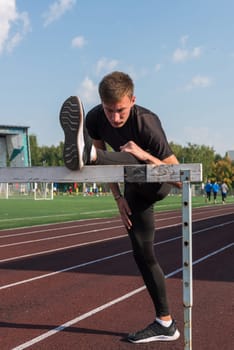 Young sporty man athlete runner in sportswear stretching before running hurdles on stadium