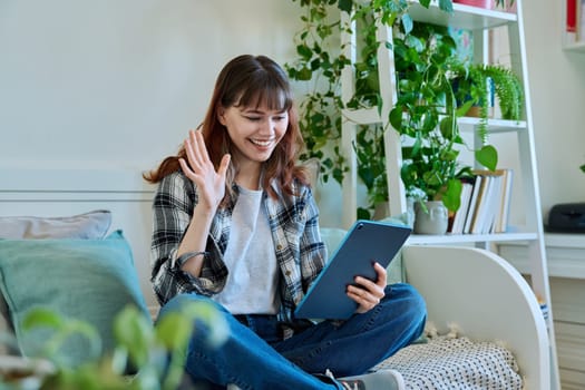 Young smiling female college student waving hand at digital tablet screen, having video chat call conference with classmates friends, sitting on couch at home. Technology, youth, education, lifestyle