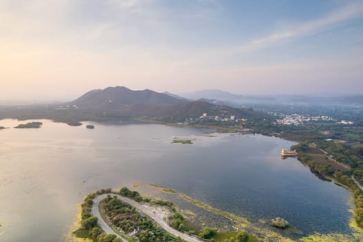 aerial drone shot showing road loop extending into fateh sagar lake with reflection of monsoon clouds with sunrise and Aravalli hills in distance in India