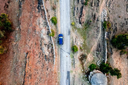 Aerial drone shot showing blue car moving on narrow country rural road with barren land on the side with green trees showing village town roads Udaipur India